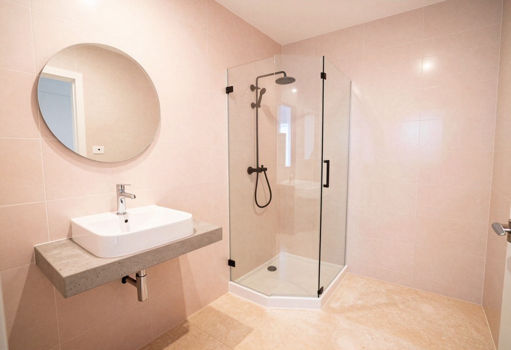Baño nuevo con mayólica rosa pálido, lavatorio sobre una repisa de concreto y ducha con mampara