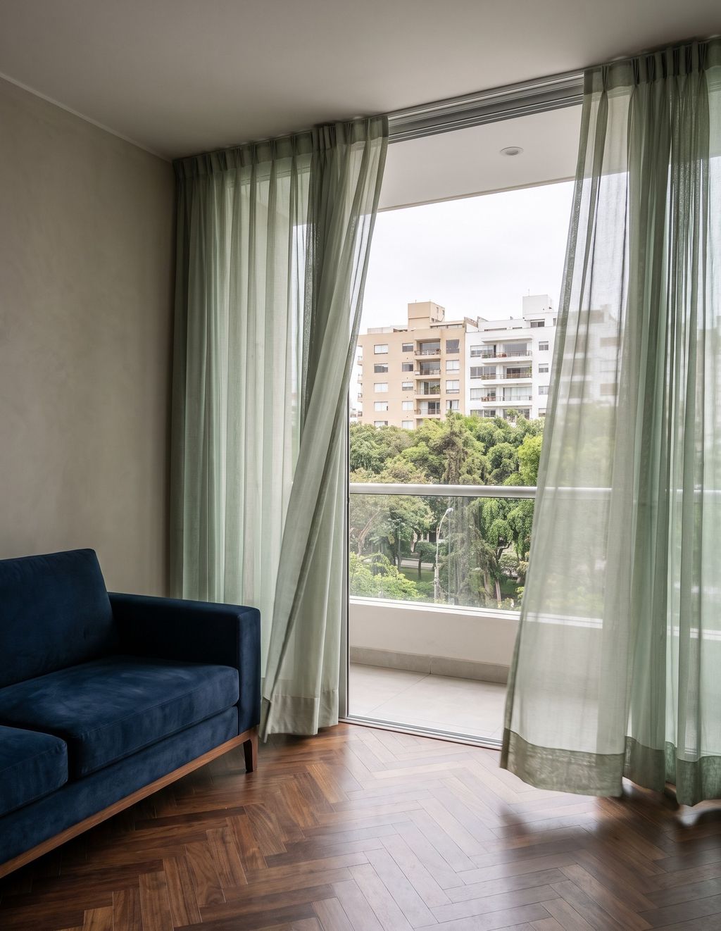 Sala de un departamento en Miraflores con cortinas de gasa verde salvia, un sofá azul de terciopelo y el balcón abierto a un parque con edificios